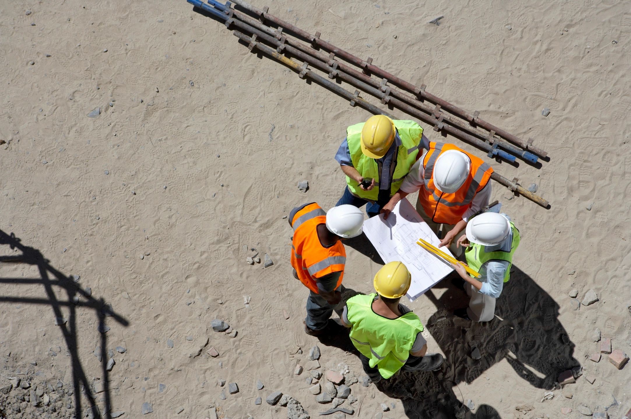 Aerial view of five construction workers gathered around a set of blueprints on a job site.